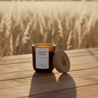 An amber glass jar candle with a cream label reading 'GOLDEN EMBER' and 'MEADOW & EMBER', shown next to its cork lid on a wheat field background placed on a wood deck.