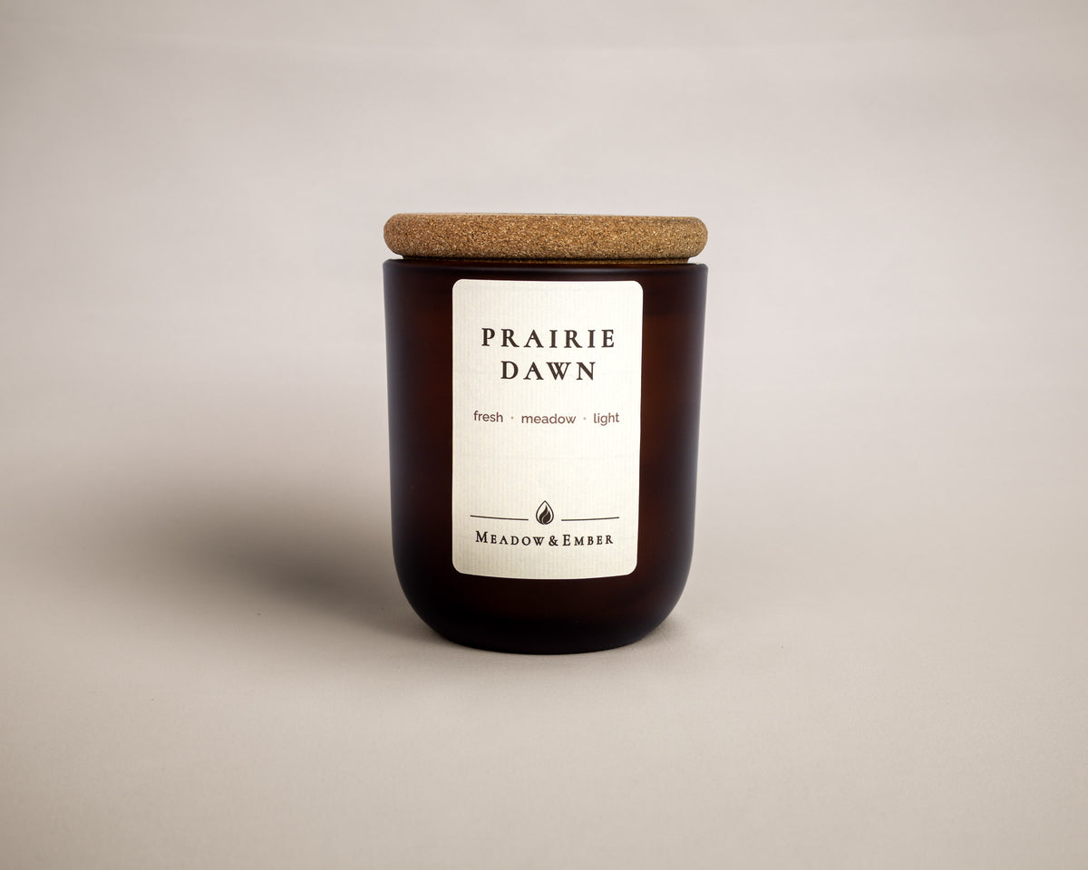 A dark amber glass jar candle with a cork lid and a white label reading 'PRAIRIE DAWN' and 'MEADOW & EMBER', centered on a light beige background.