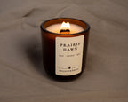 A lit amber glass jar candle with a white label reading 'PRAIRIE DAWN' and 'MEADOW & EMBER' on a textured taupe fabric background.