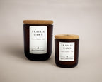 Two amber glass jar candles with cork lids and white labels reading PRAIRIE DAWN by Meadow & Ember, shown on a warm beige studio background.