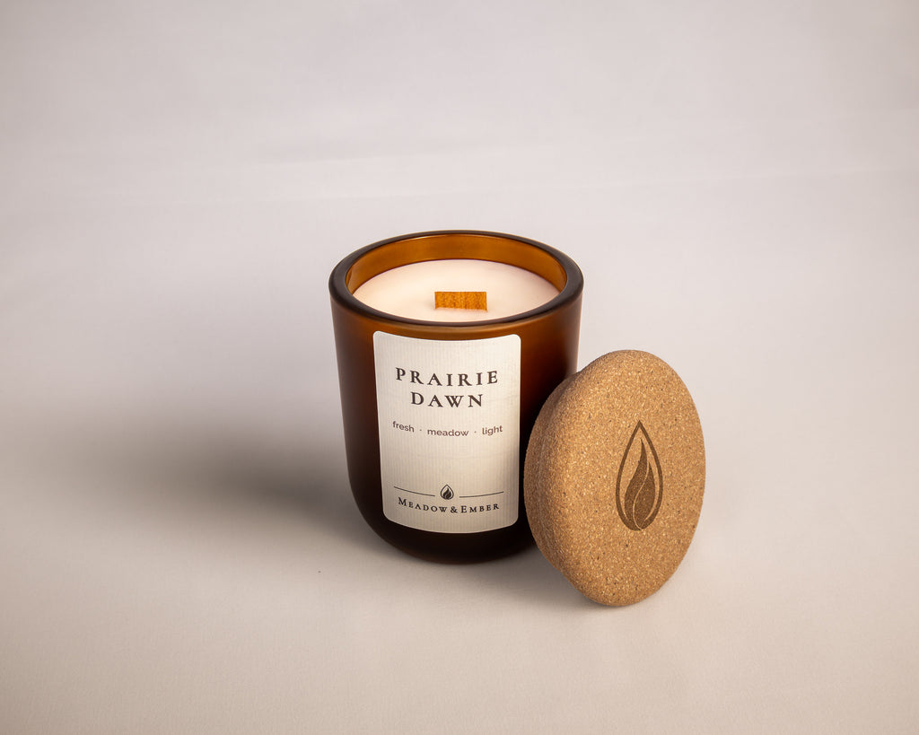 An amber glass jar candle with a wood wick labeled 'Prairie Dawn' by Meadow & Ember, shown with its cork lid leaning against it on a light neutral background.