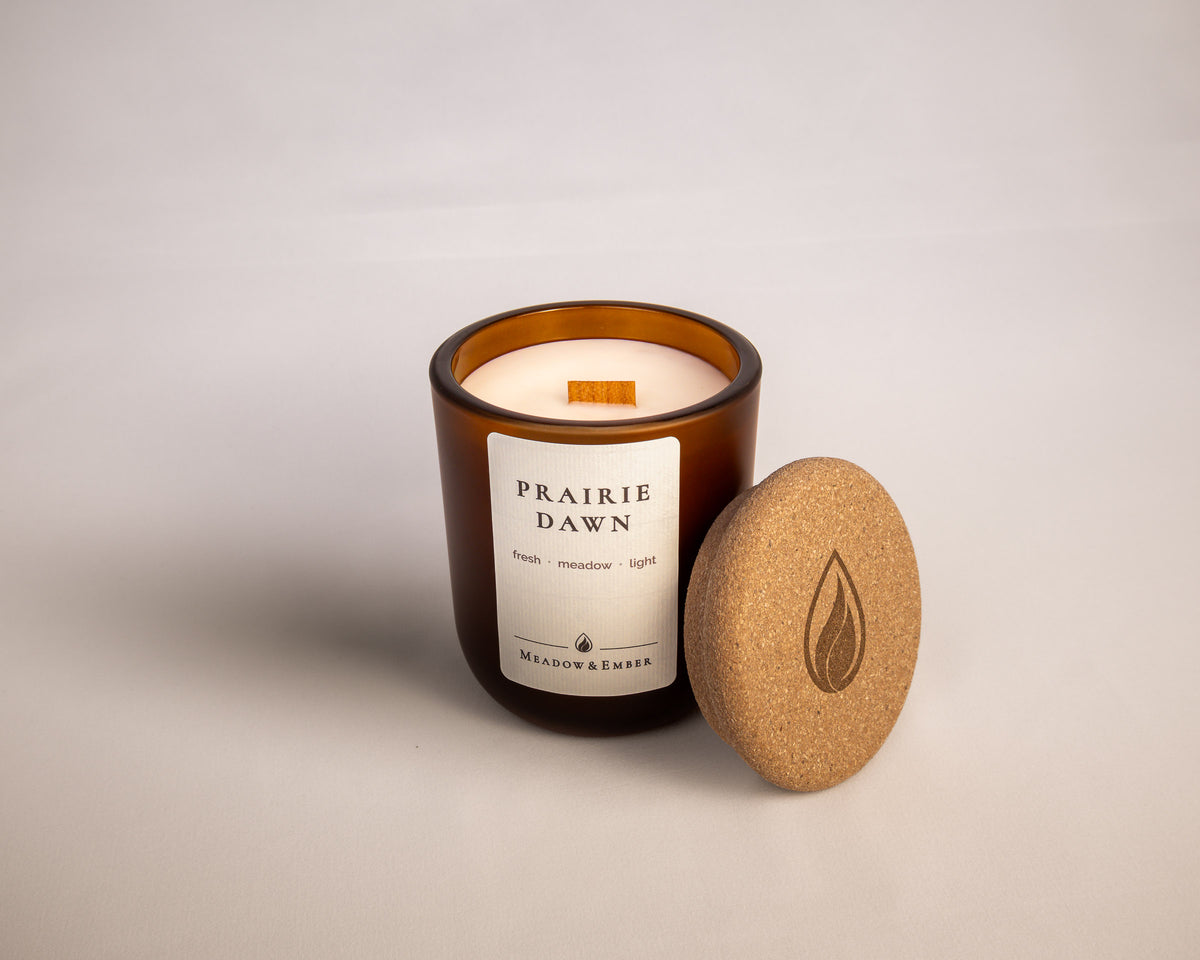 An amber glass jar candle with a wood wick labeled 'Prairie Dawn' by Meadow & Ember, shown with its cork lid leaning against it on a light neutral background.